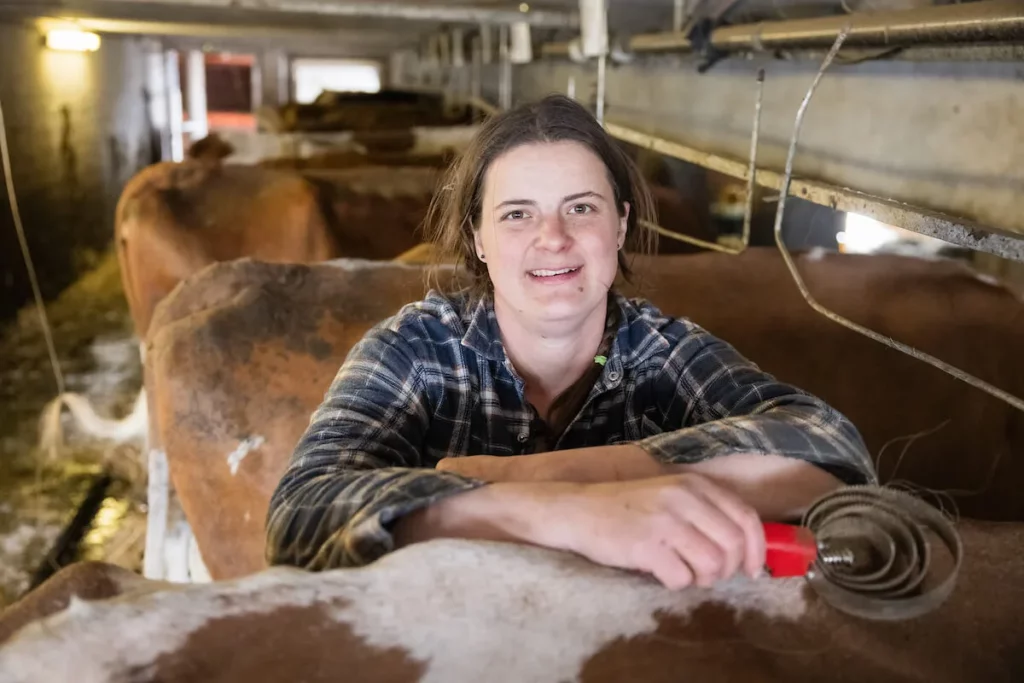 Femme agricultrice souriante appuyée sur une barrière dans une étable, entourée de vaches.
